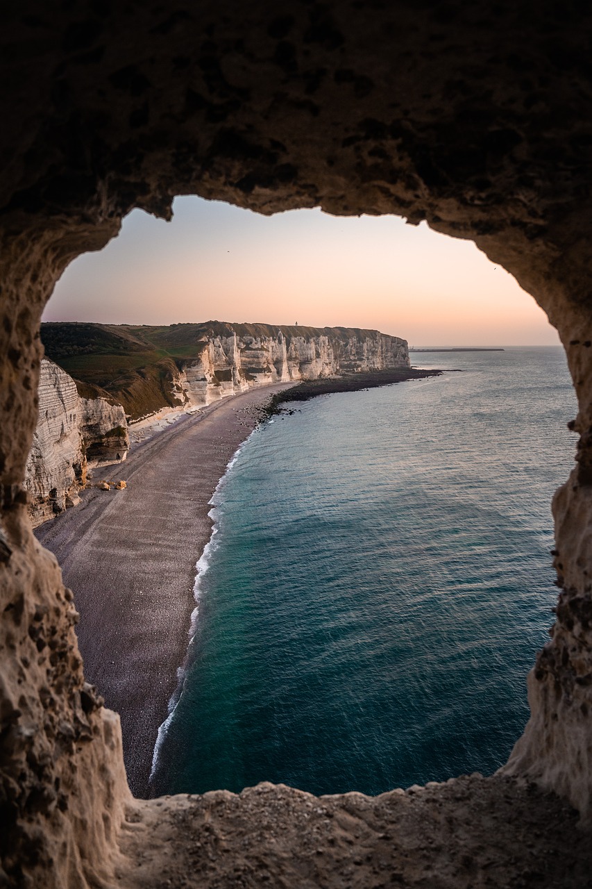 En la imagen se ve, desde una cueva el amanecer de un nuevo día en una costa, frente al mar, con una playa desierta y sin oleaje, un día calmado.